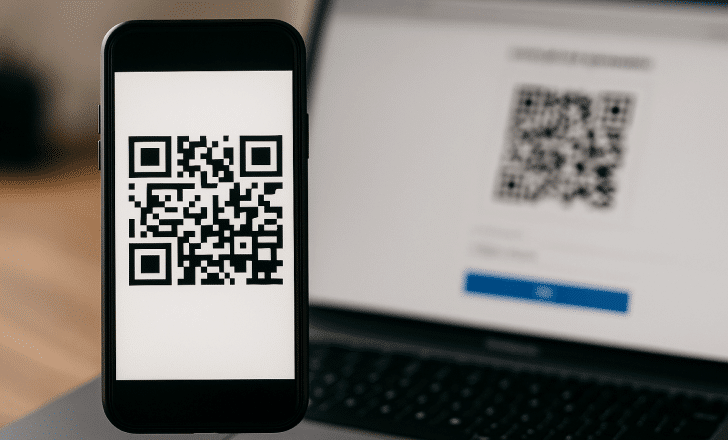A smartphone displaying a QR code, with a blurred laptop in the background showing a QR code generator interface