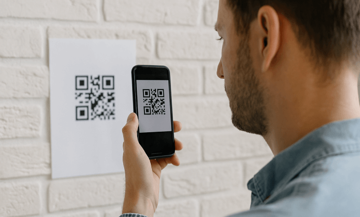A man scanning a QR code printed on a sheet of paper taped to a white brick wall