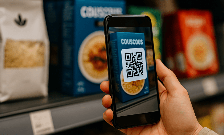 A person holds a modern smartphone in front of a blue couscous box on a grocery store shelf, displaying a black-and-white QR code aligned with the packaging design.